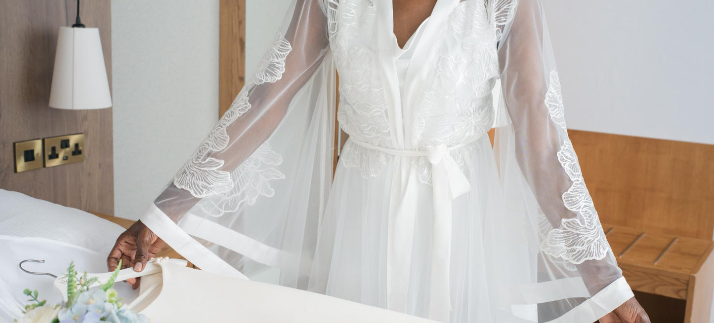 Woman in a white bridal robe standing in front of a mirror, holding a bouquet of flowers.