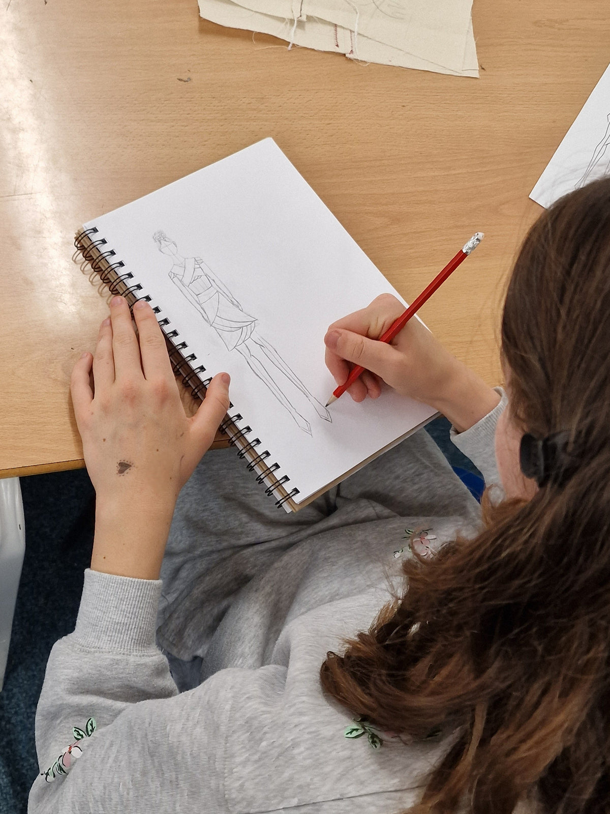 A person with long brown hair sits at a wooden desk, drawing a figure in a spiral notebook with a red pencil.